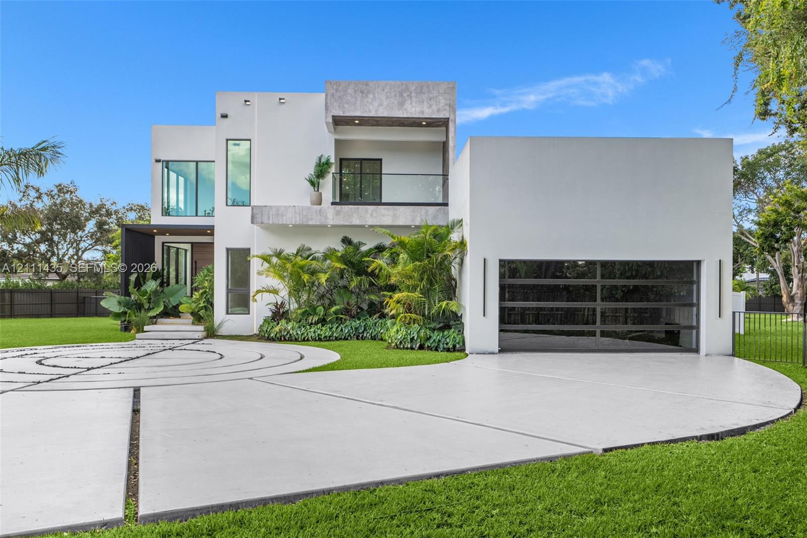 Modern two-story exterior featuring smooth stucco siding, a second-level balcony with glass railing, tiered concrete driveway with decorative inlay, and an oversized multi-panel garage door