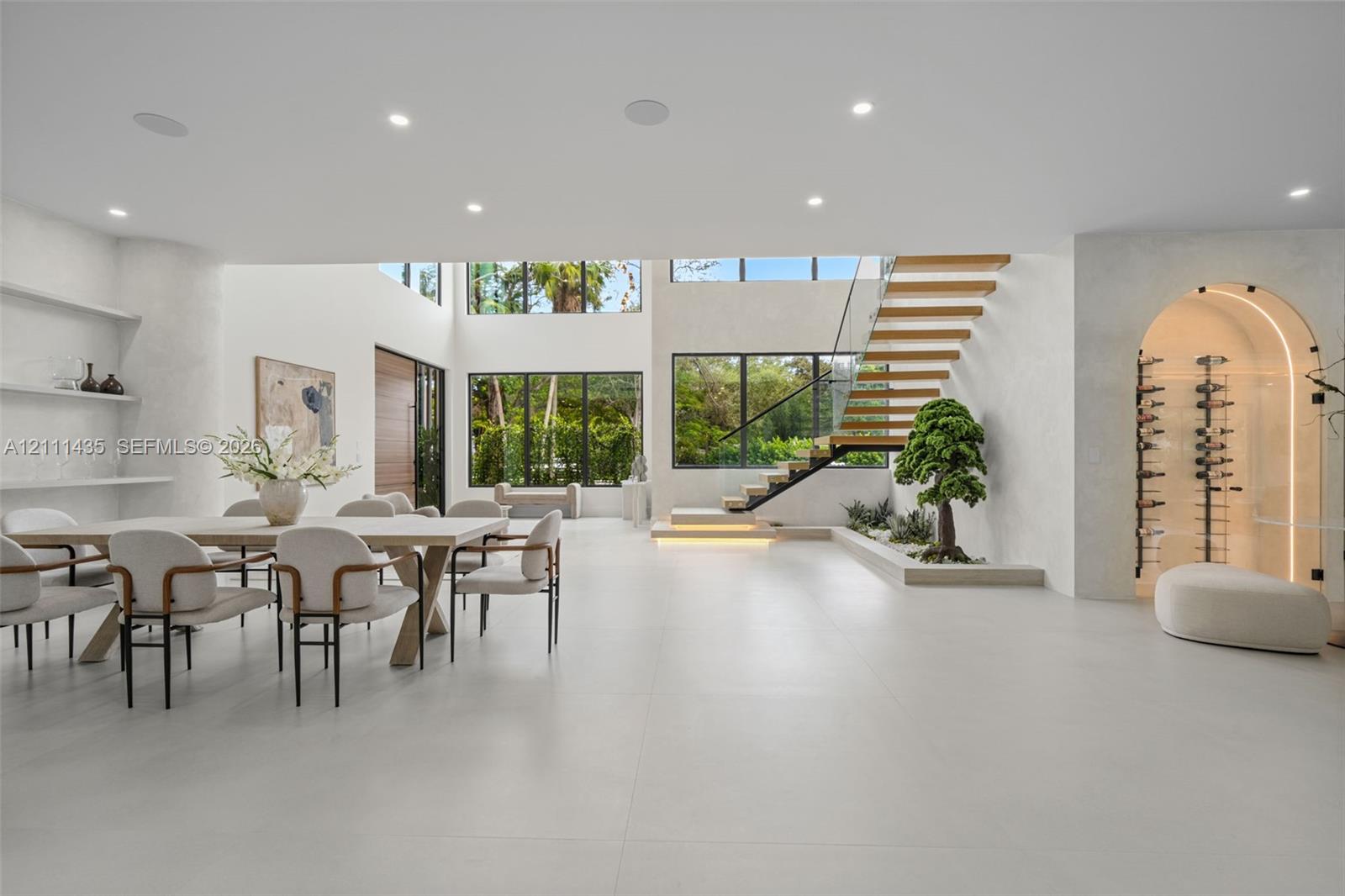 Open-concept living space featuring large-format tile flooring, a floating staircase with glass railings, built-in recessed shelving, a glass-enclosed arched wine display, and expansive floor-to-ceiling windows