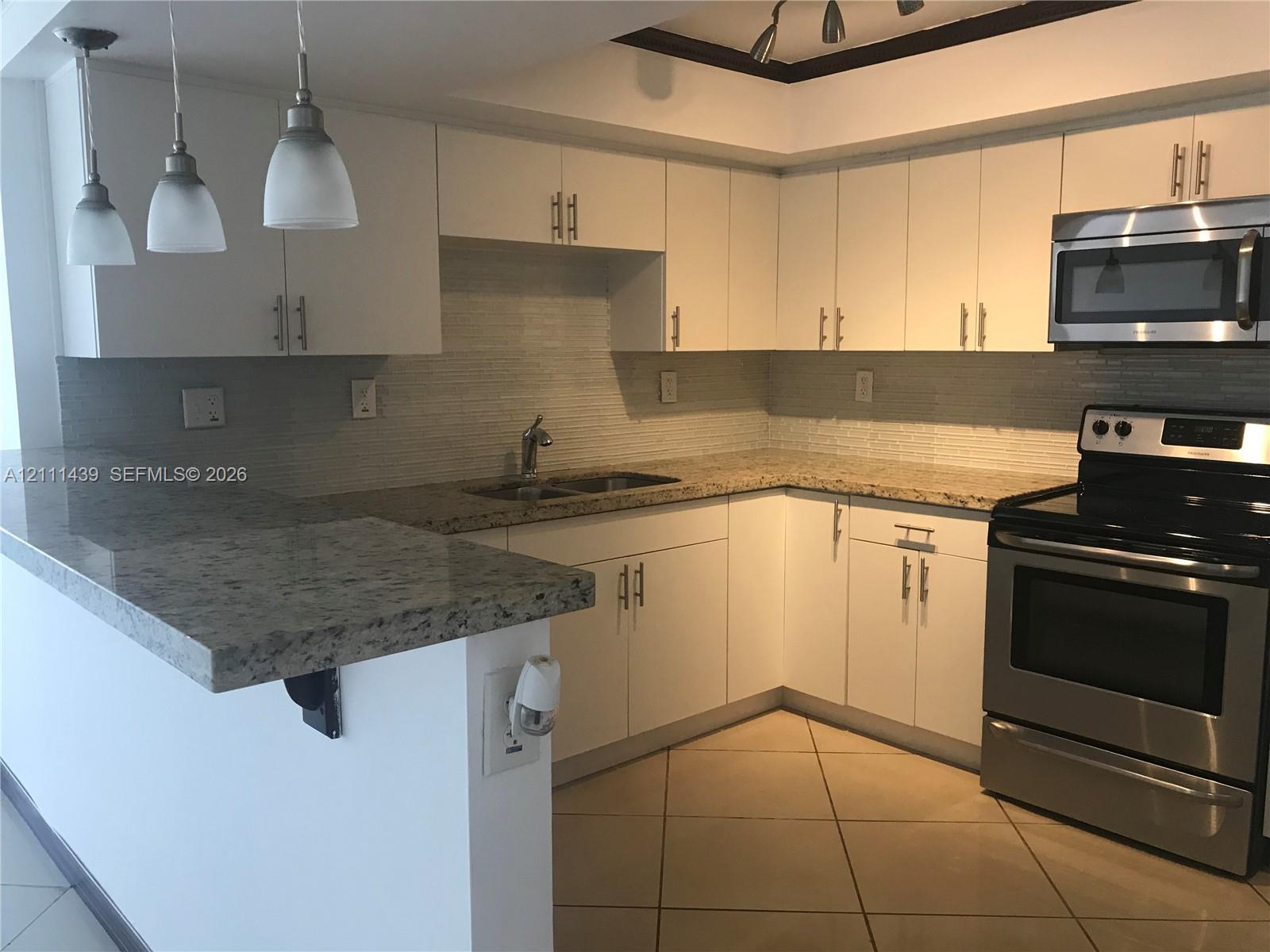 Kitchen featuring white cabinetry with brushed metal hardware, stone-surface countertops, stainless steel appliances, a tiled backsplash, and three pendant lights