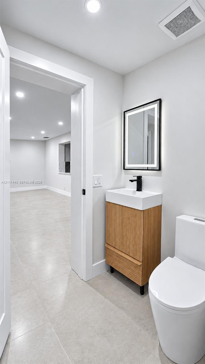 Modern vanity with wood-finish cabinetry, white basin, matte black faucet, and rectangular lighted mirror, complemented by ceramic tile flooring and a minimalist dual-flush toilet