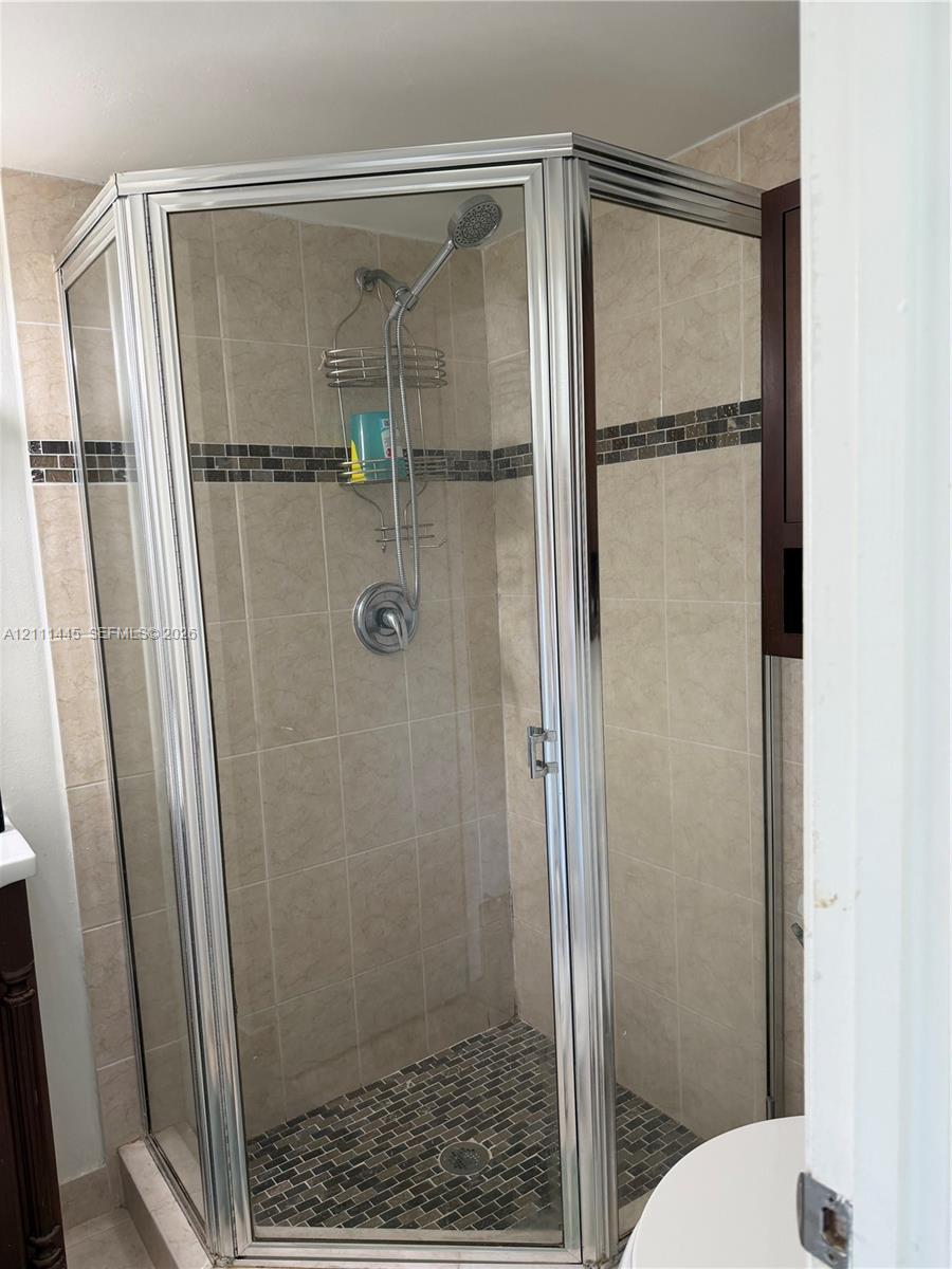 Corner shower enclosure featuring a metal frame, tile wall surround, stone-tiled floor, decorative glass tile accent strip, and a chrome hand-held shower fixture