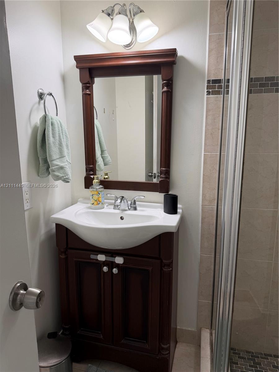 Wood-finish vanity with an integrated white sink, coordinated wood-frame mirror, three-light polished chrome fixture, and tiled shower stall with a decorative mosaic glass accent border