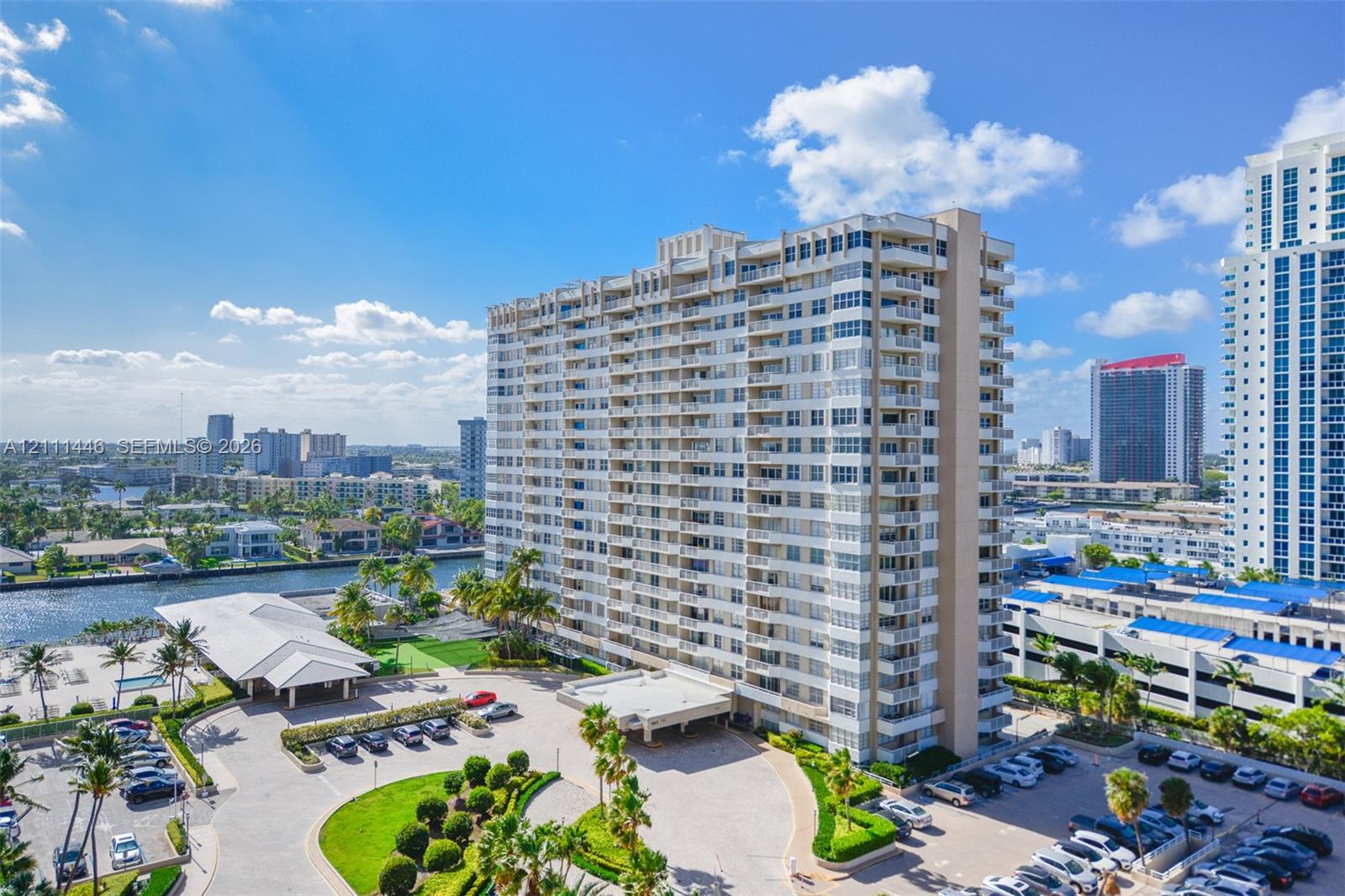 High-rise residential building featuring stacked private balconies, an expansive paved motor court with covered entrance, manicured landscaping, and water frontage