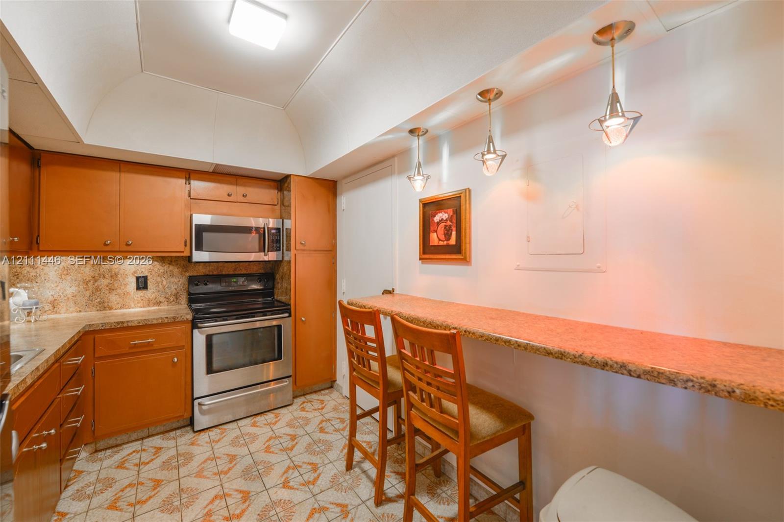 Kitchen featuring wood cabinetry, stainless steel appliances, speckled stone-finish countertops, tile flooring, and three pendant lights over a built-in breakfast bar