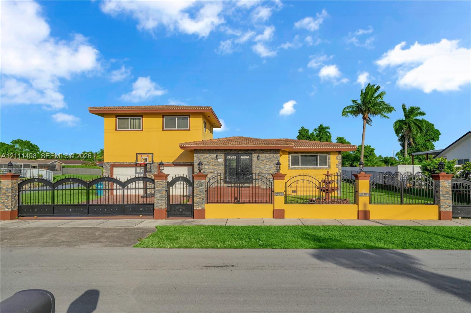 Exterior featuring a two-story stucco facade, decorative stone accents, a multi-tiered fountain, and perimeter wall with wrought iron fencing