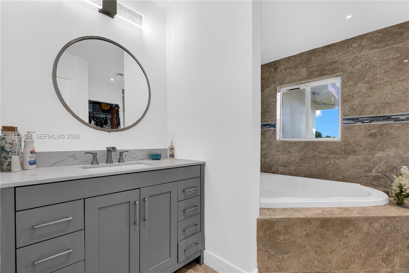 Gray cabinetry vanity an integrated rectangular sink, a circular mirror with metallic framing, and a tiled soaking tub