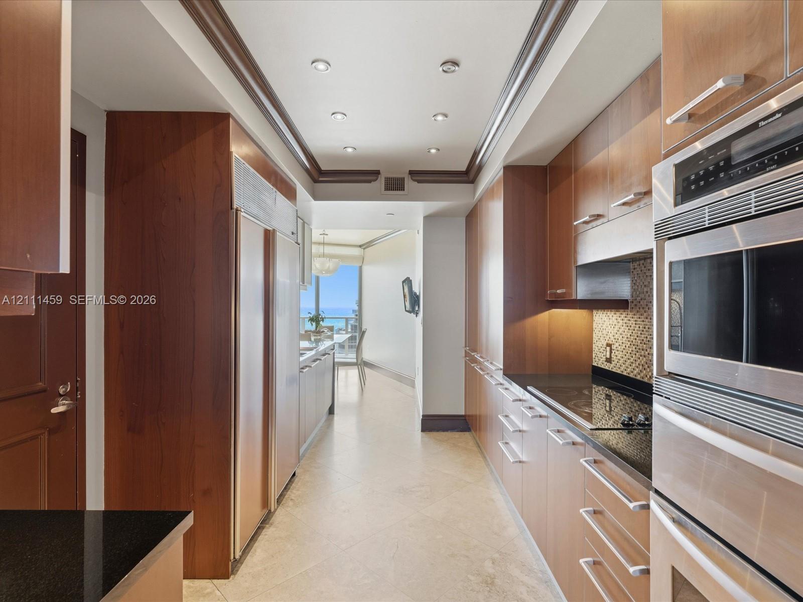 Galley kitchen featuring custom flat-panel wood cabinetry, black stone countertops, integrated stainless steel refrigeration, built-in double ovens, and recessed ceiling lighting