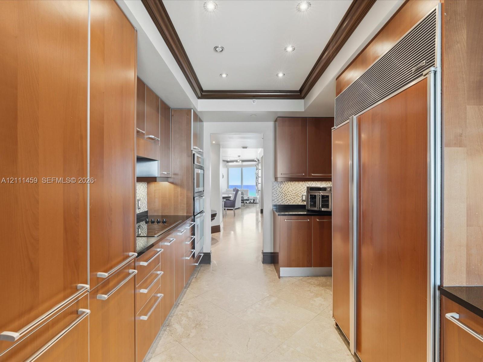 Galley kitchen featuring custom flat-panel wood cabinetry, integrated stainless steel refrigerator, black stone countertops, tile flooring, and recessed ceiling lighting with crown molding