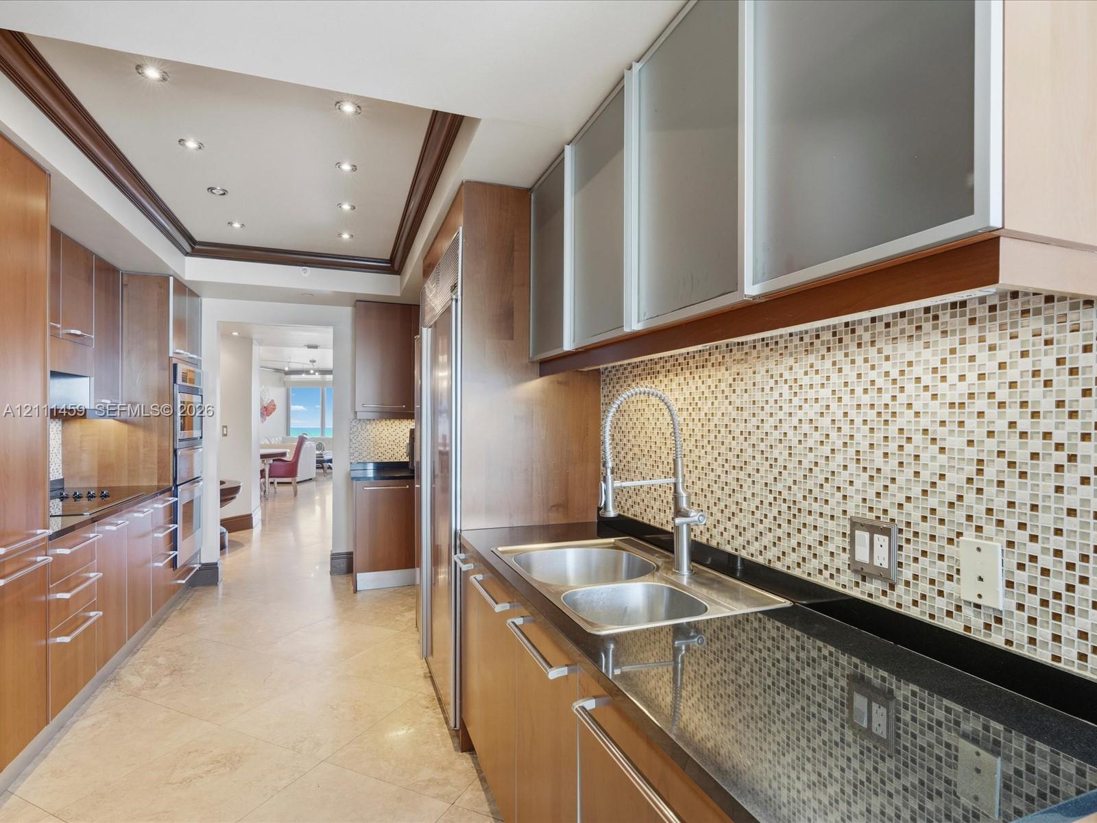 Kitchen featuring wood cabinetry, frosted-glass upper cabinet doors, dark solid-surface countertops, mosaic tile backsplash, and stainless steel double-basin sink with pull-down faucet