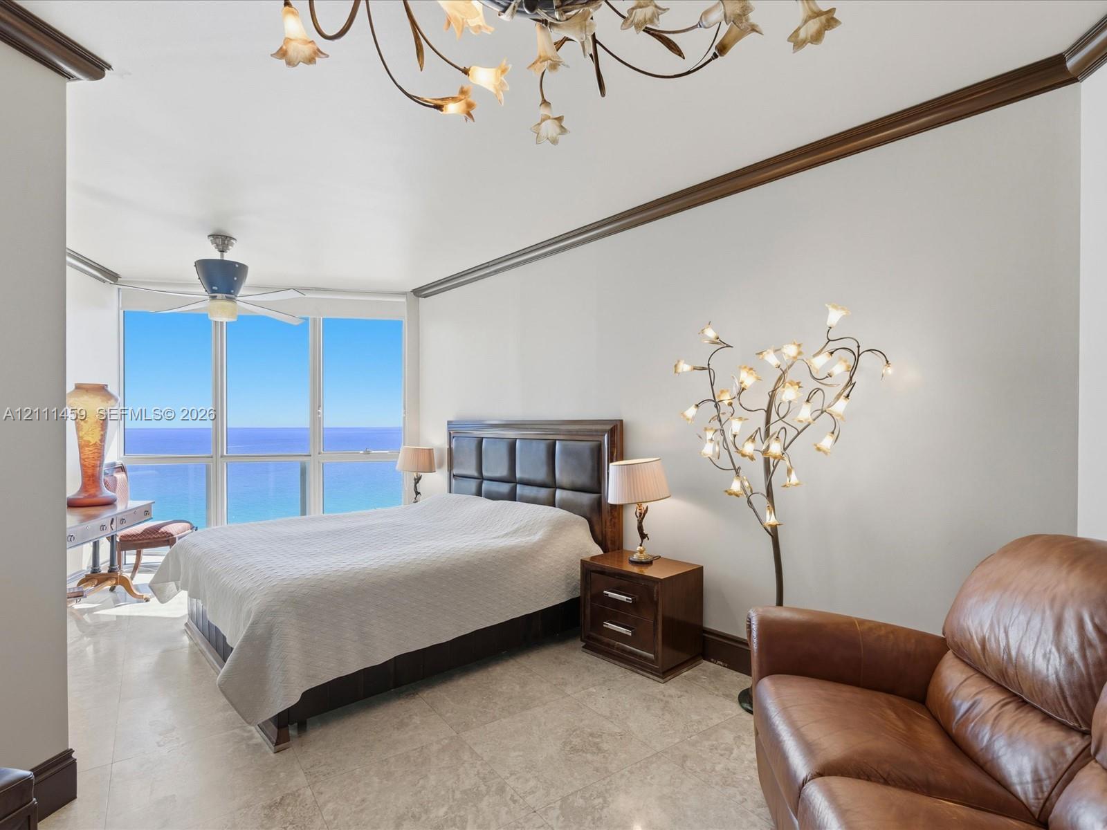 Floor-to-ceiling windows featuring ocean horizon, neutral stone flooring, decorative crown molding, and intricate multi-light chandelier