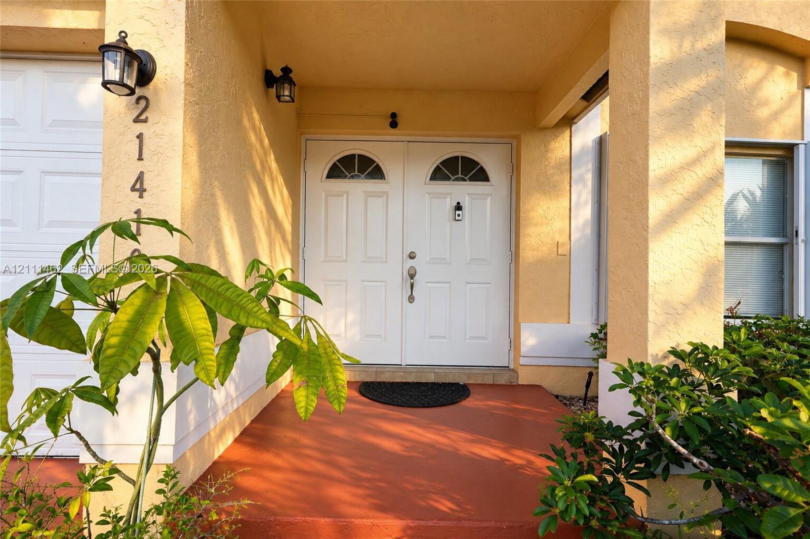 Covered entryway featuring double doors with arched glass inserts, concrete porch flooring, stucco exterior, and wall-mounted coach lighting