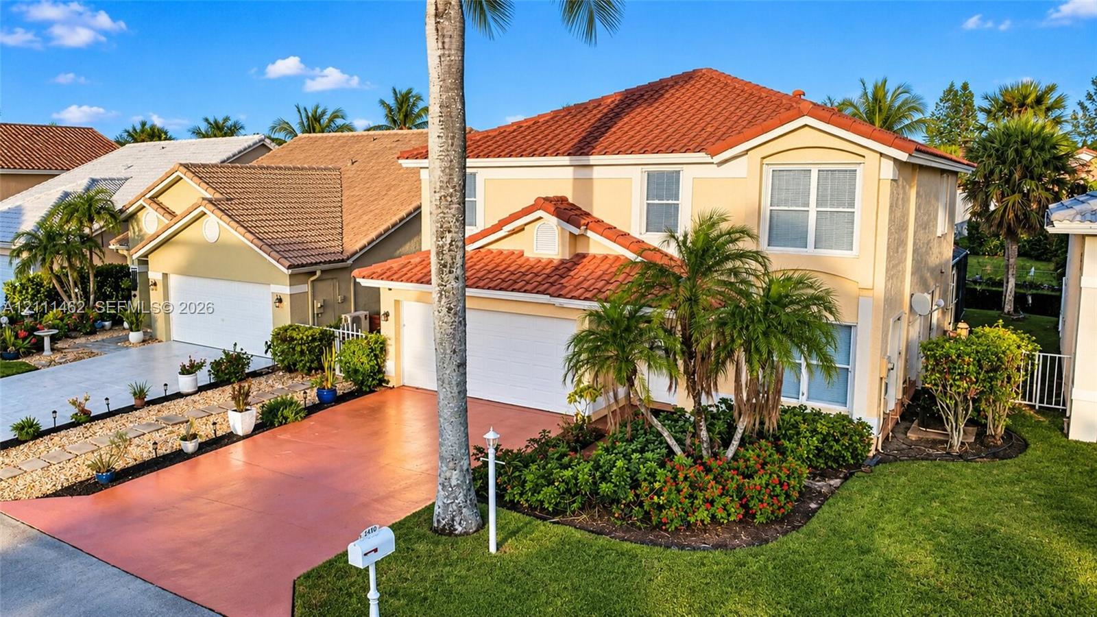 Two-story residence featuring a terracotta-tiled roof, stucco exterior, two-car garage, painted concrete driveway, and mature landscaping Two-story residence featuring a terracotta-tiled roof, stucco exterior, two-car garage, painted concrete driveway, and mature landscaping
