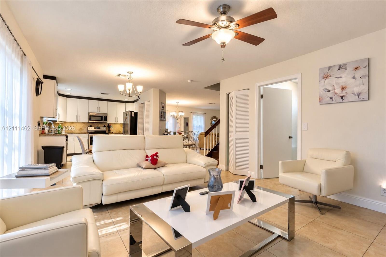 Open-concept living space featuring ceramic tile flooring, a multi-blade ceiling fan with integrated lighting, shaker-style kitchen cabinetry, and louvered-panel closet doors