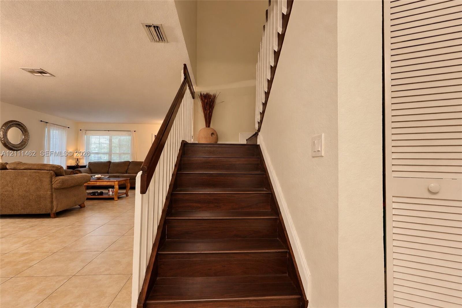 Staircase featuring dark wood-finish treads, white balusters, and a dark wood handrail, paired with neutral floor tiling and louvered closet doors