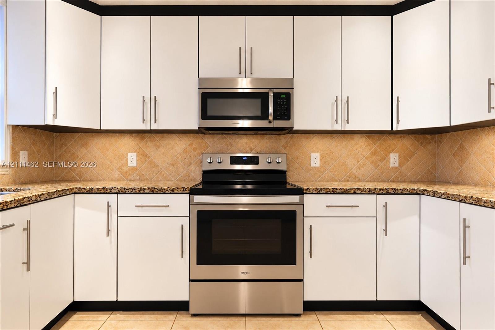 White shaker-style cabinetry with modern hardware, stone-surface countertops, neutral diagonal-set tile backsplash, and stainless steel appliances including a built-in microwave and range