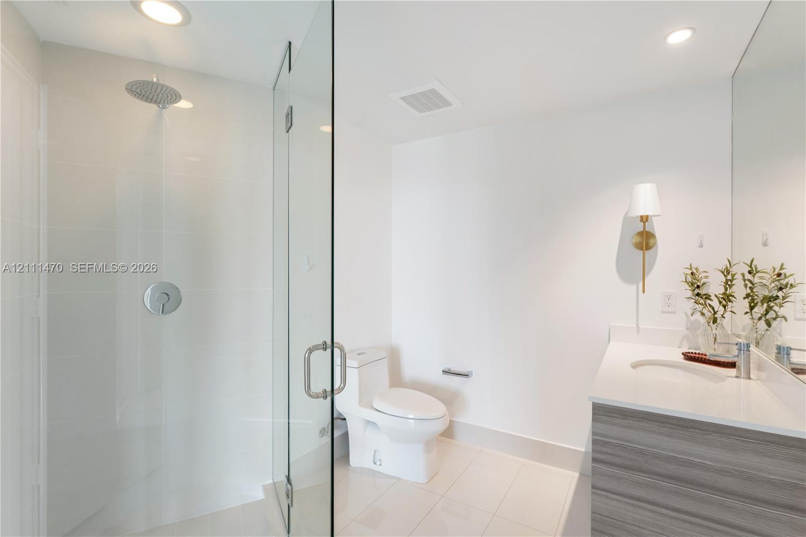 Walk-in shower with glass enclosure and rainfall showerhead, vanity with stone countertop, wood-finish cabinetry, and wall-mounted lighting