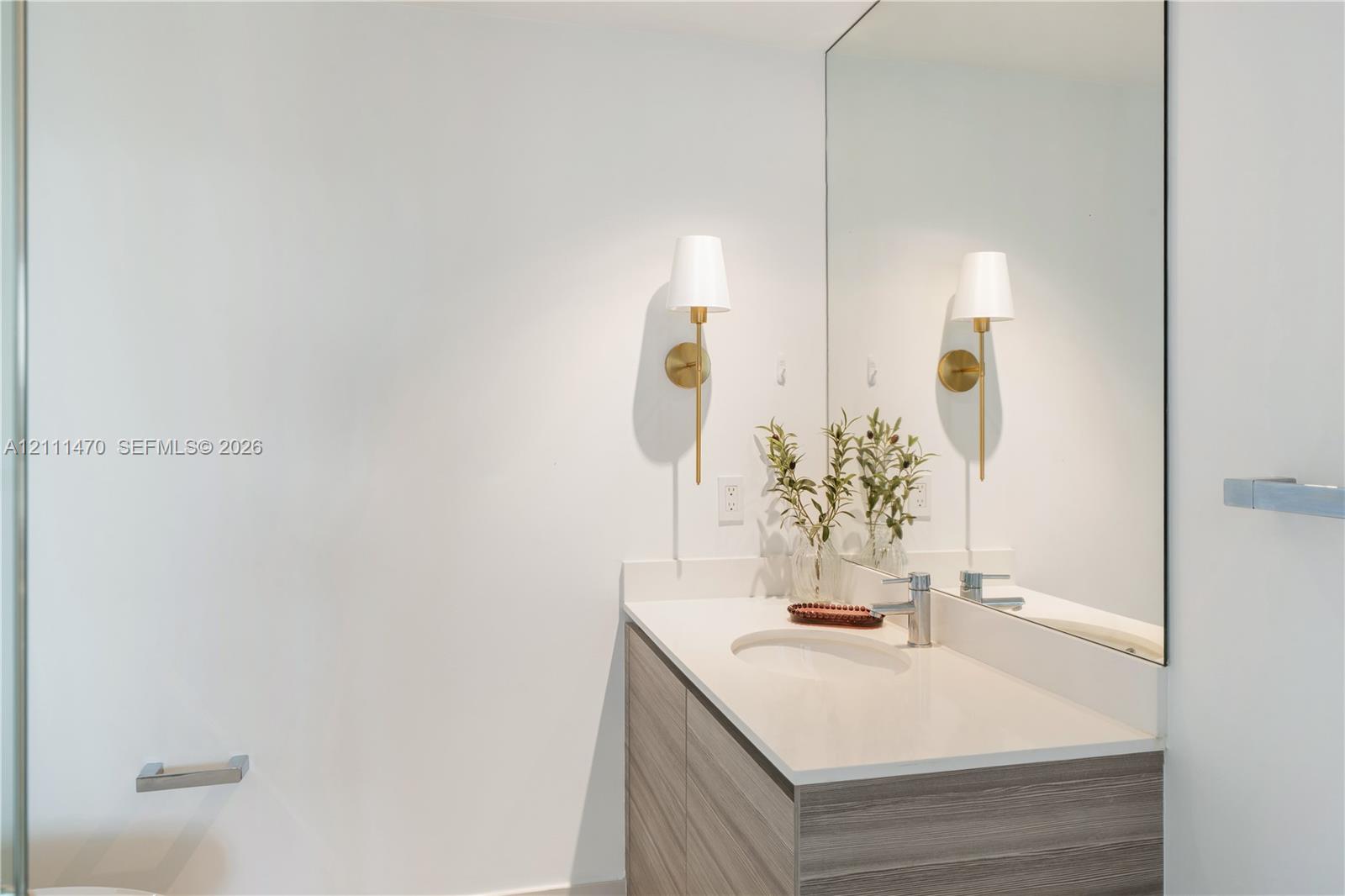 Modern vanity featuring a stone-composite countertop with an integrated oval sink, horizontal wood-finish cabinetry, a large frameless wall mirror, and dual brass-toned wall sconces