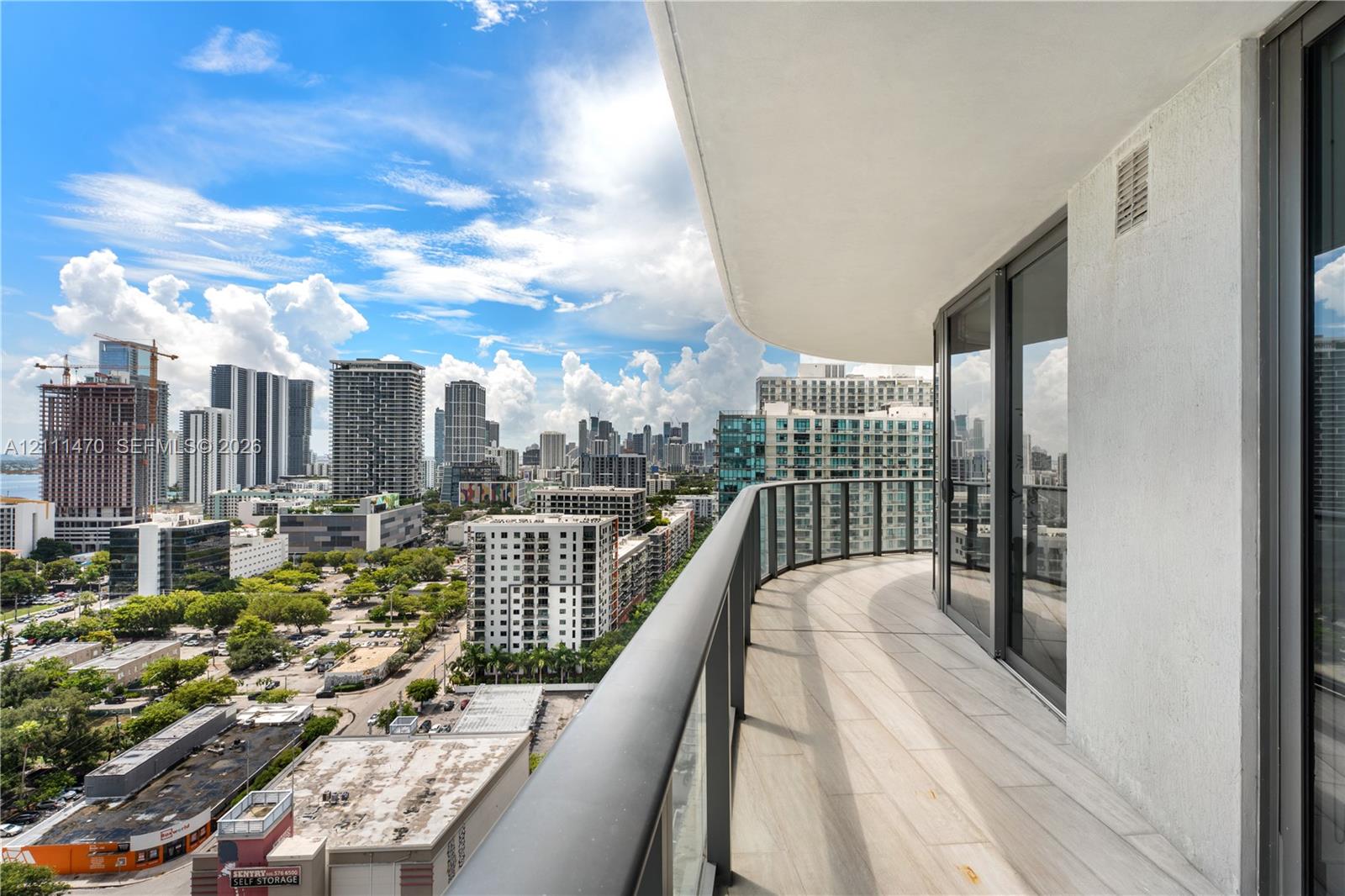 Curved balcony featuring light-toned tile flooring, glass railings with a dark metal frame, and sliding glass entry doors