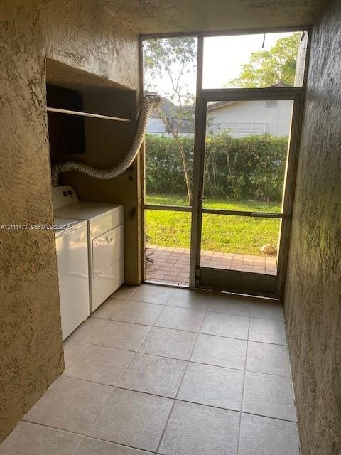Laundry area featuring a built-in recessed shelf, light-colored tile flooring, stucco walls, and a screen door with exterior access to a paved patio area