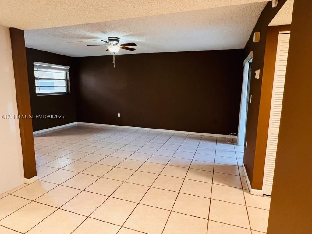 Open-concept interior featuring light-toned tile flooring, a ceiling fan with integrated light fixture, recessed window with horizontal blinds, and louvered closet doors