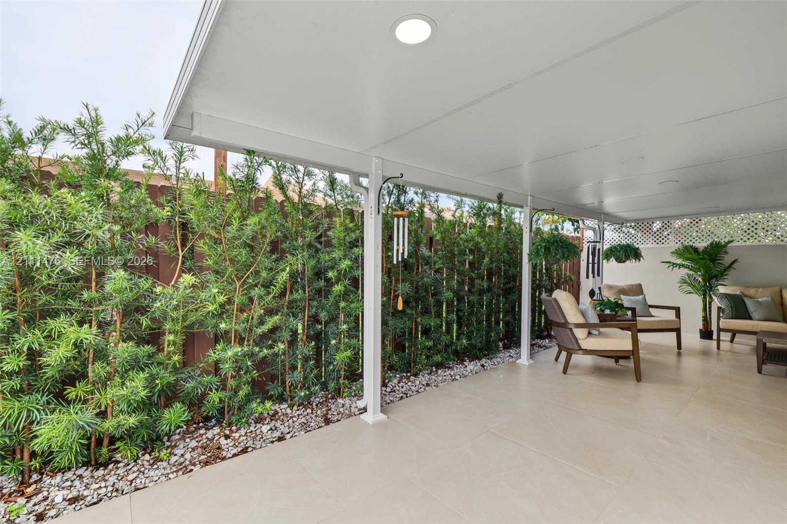 Covered patio featuring large-format tile flooring, recessed lighting, and a perimeter of mature landscaping set against a privacy fence