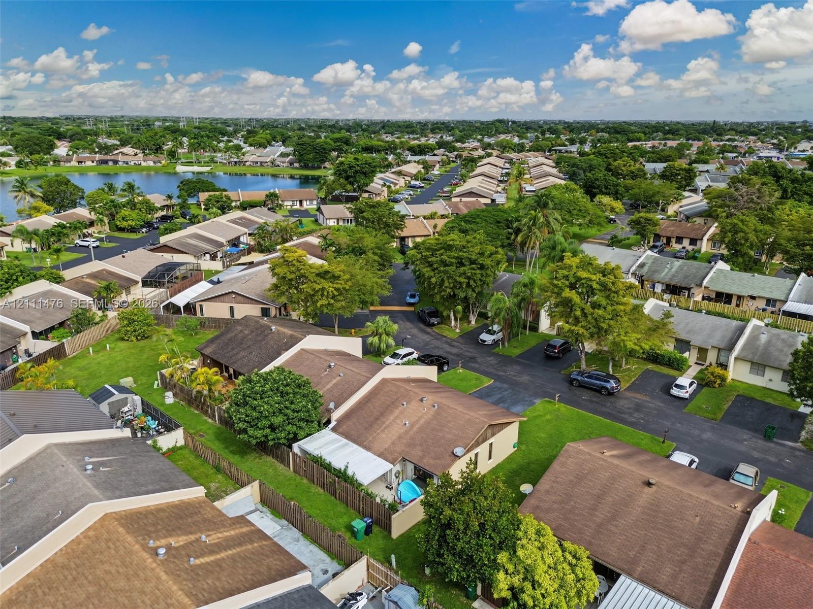 Aerial perspective of a residential community featuring shingle-roofed structures, expansive green space, mature trees, asphalt roadways, and a lake