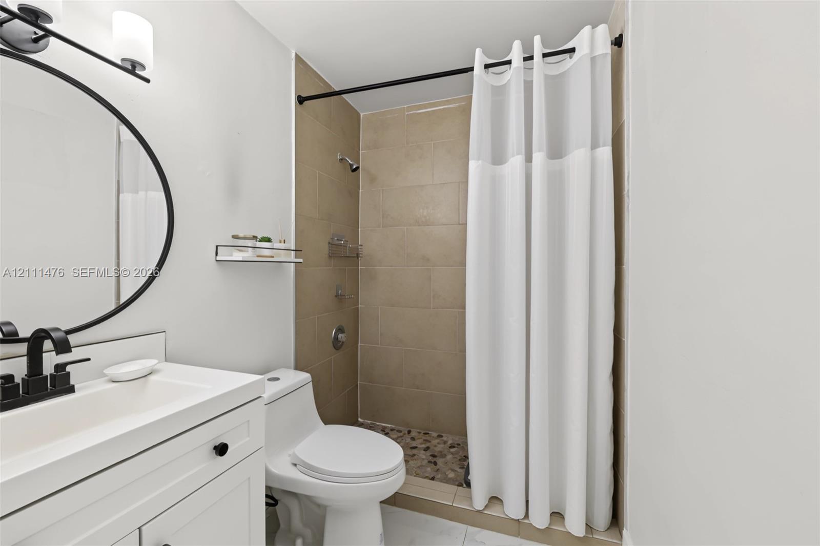 White vanity with integrated rectangular sink and matte black faucet, round wall-mounted mirror, matte black vanity lighting, shower enclosure with neutral-toned wall tiles, and stone-tiled shower flooring