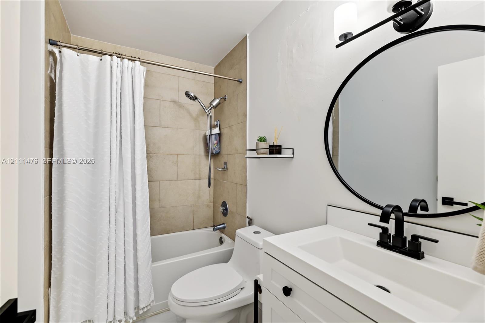 Bathroom featuring a white vanity with an integrated basin, matte black widespread faucet, circular mirror with a black frame, wall-mounted vanity lighting, and a bathtub with beige tiled surround