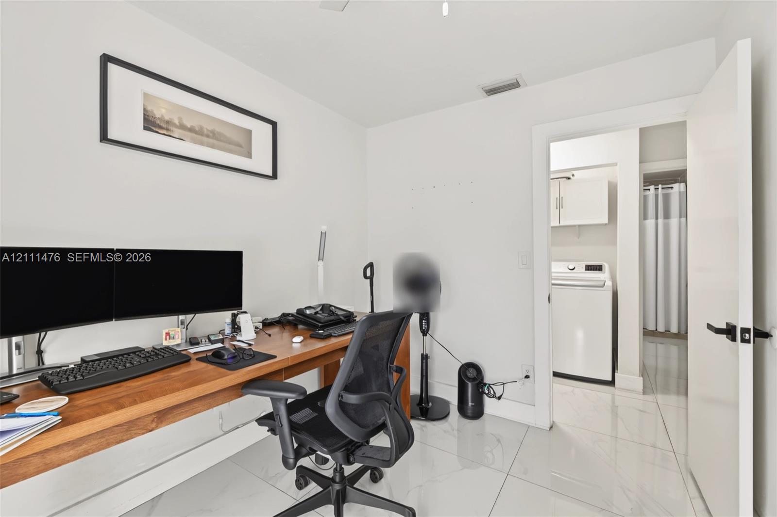 Polished stone-finish tile flooring, built-in natural wood desk, and interior doorway providing access to a laundry area with overhead cabinetry