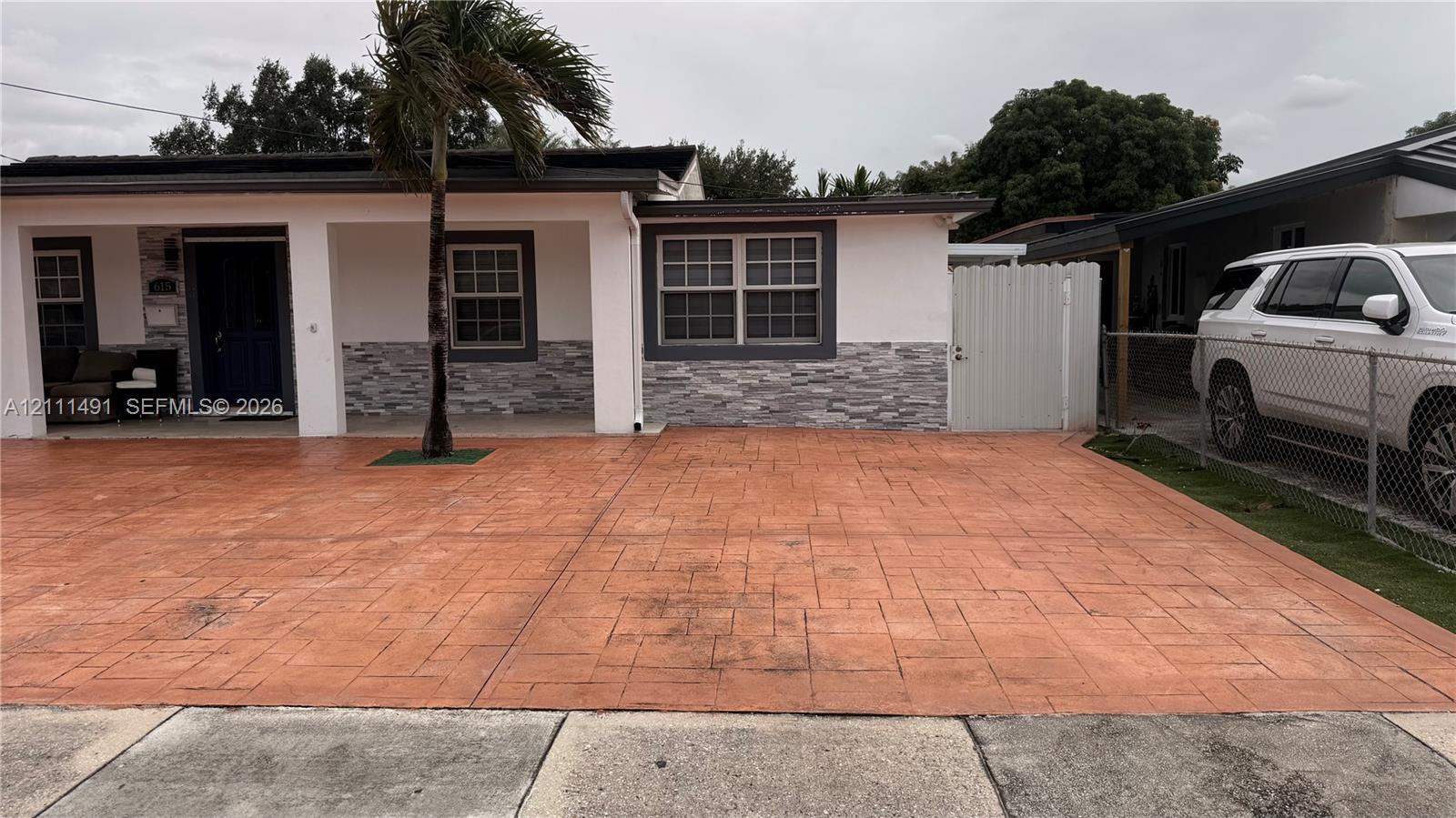 Exterior facade featuring stone veneer detailing, a covered entryway, stamped concrete driveway, and white privacy fencing Exterior facade featuring stone veneer detailing, a covered entryway, stamped concrete driveway, and white privacy fencing