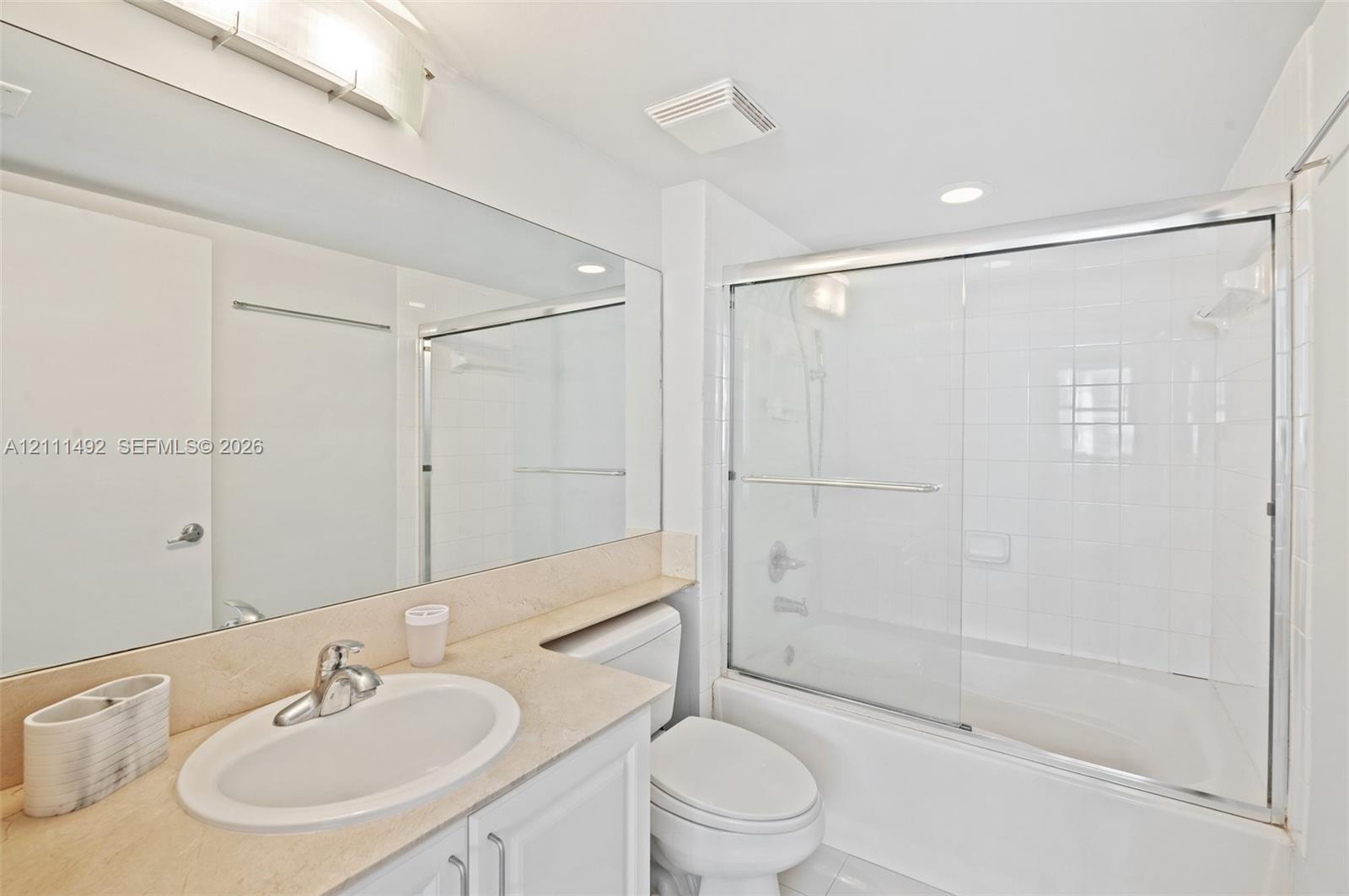 Full-width vanity with solid-surface countertop and integrated oval sink, expansive wall-mounted mirror, white cabinetry, and shower-tub combination featuring a sliding glass enclosure and full-height tiled surround