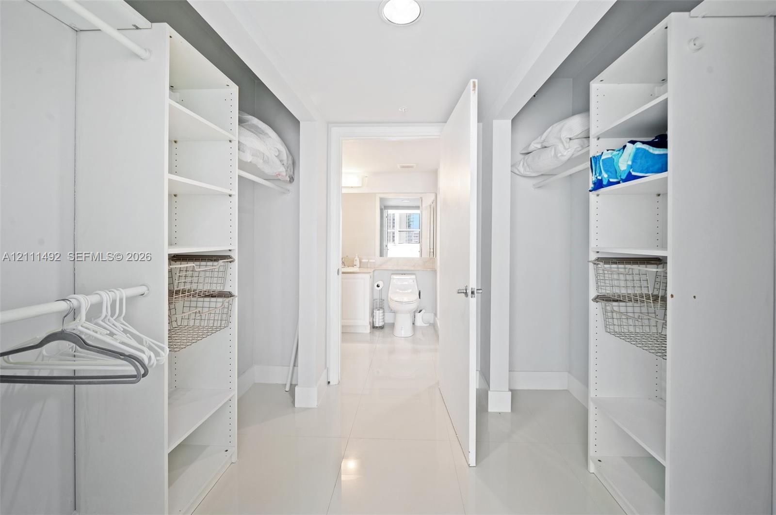 Walk-in closet featuring floor-to-ceiling white built-in shelving, integrated metal basket organizers, dual hanging rods, and high-gloss tile flooring