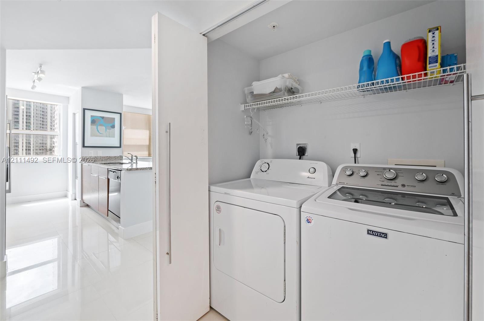Laundry closet featuring a wire shelving unit and stacked washer and dryer, polished tile flooring, and a sleek kitchen space with stone-surfaced countertops and stainless steel appliances
