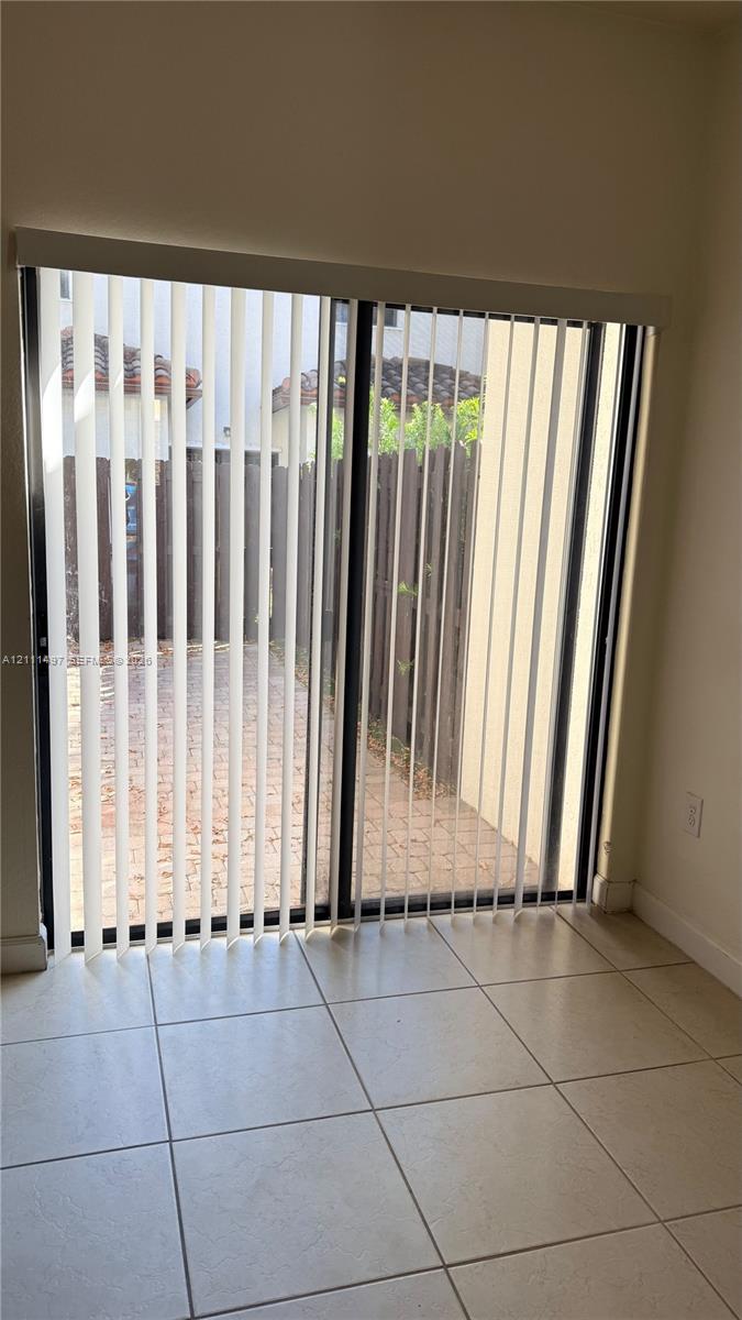 Sliding glass door with vertical blinds, neutral tile flooring, and a paved outdoor patio area enclosed by a wooden fence