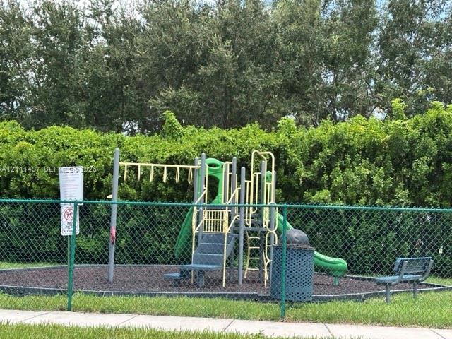 Playground area featuring a multi-component play structure with a slide and climbing features, enclosed by green chain-link fencing, surrounded by mature perimeter hedging, and incorporating a permanent waste receptacle and stationary bench