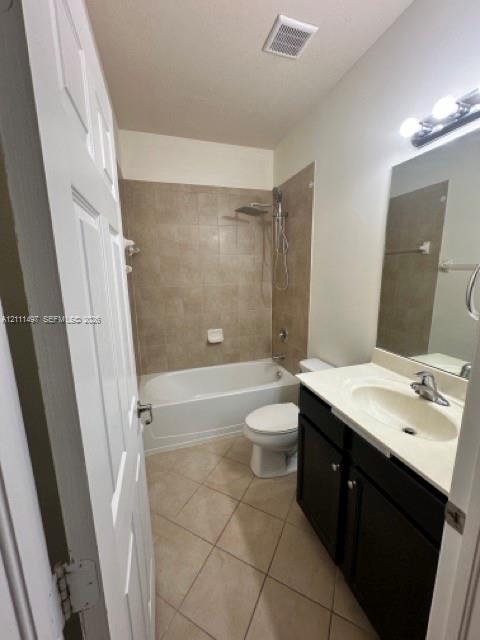 Bathroom featuring a vanity with dark cabinetry and a light-toned countertop, a shower-tub combination with full-height tile surround, wall-mounted light fixture, and diagonal tile flooring
