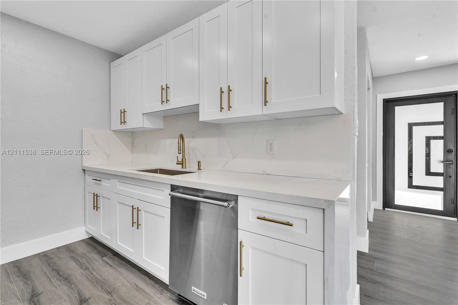 Kitchen featuring white Shaker-style cabinetry, gold-tone hardware, stone-finish countertops with matching backsplash, stainless steel dishwasher, and wood-finish flooring