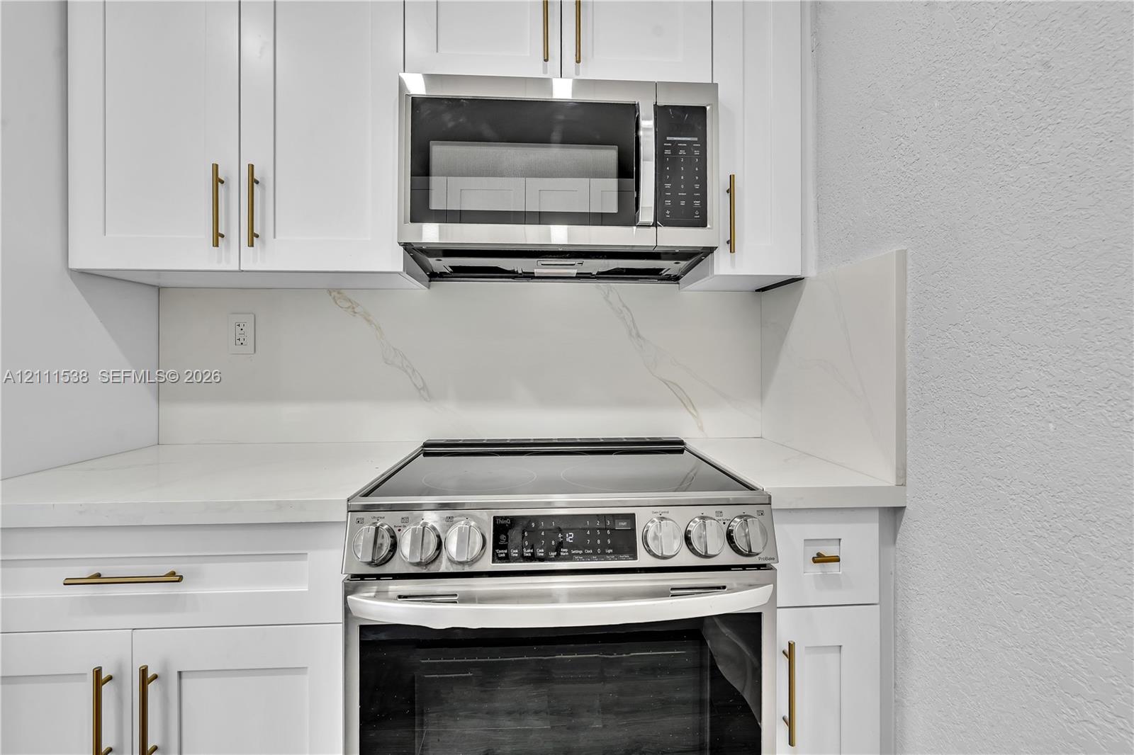Kitchen featuring white shaker cabinetry with gold-toned hardware, stone-composite countertops, integrated stone backsplash, stainless steel electric range, and matching stainless steel microwave