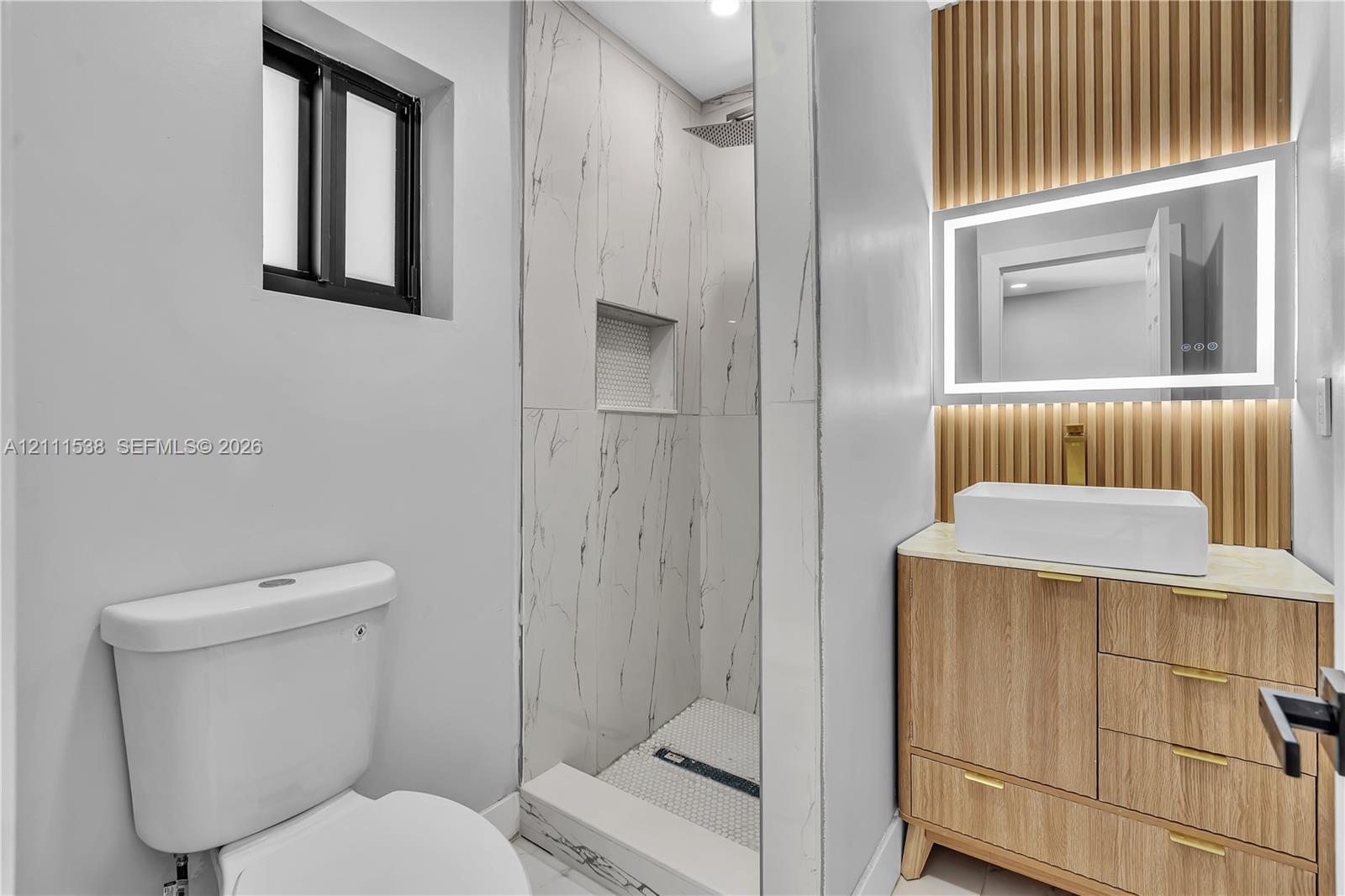 Bathroom featuring a wood-finish vanity with a rectangular vessel sink, LED-backlit mirror, vertical wood-slat accent wall, and a walk-in shower with large-format tile surrounds and a built-in niche