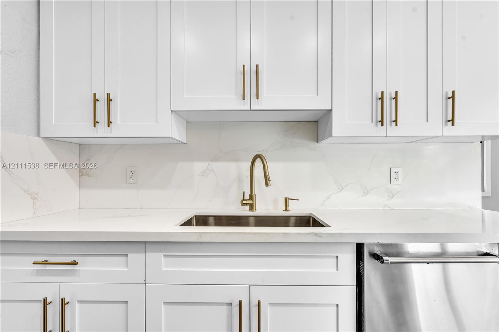 Shaker-style white cabinetry with metallic pulls, white stone-finish countertops, full-height stone-finish backsplash, undermount stainless steel sink with a high-arc metallic faucet, and stainless steel dishwasher
