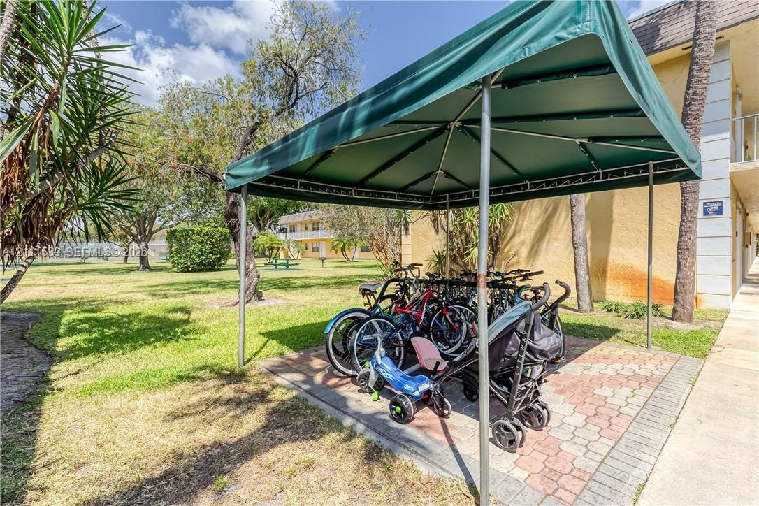 Covered outdoor storage area with a metal frame and fabric canopy situated on a paved brick foundation, set within an expansive grass-covered grounds area featuring mature trees