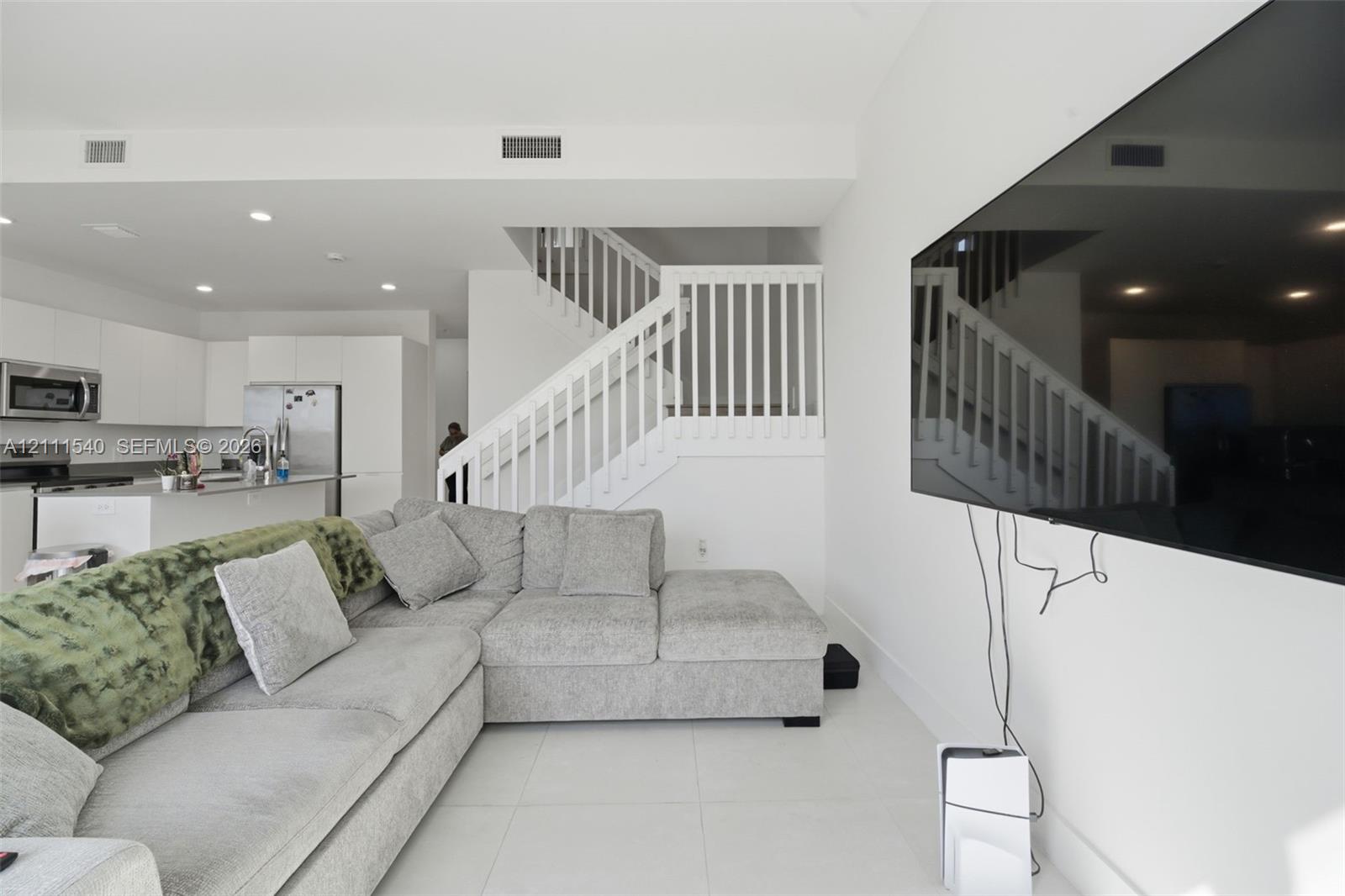 Open-concept interior featuring light-toned tile flooring, recessed ceiling lighting, a staircase with white balusters, and a kitchen equipped with white cabinetry