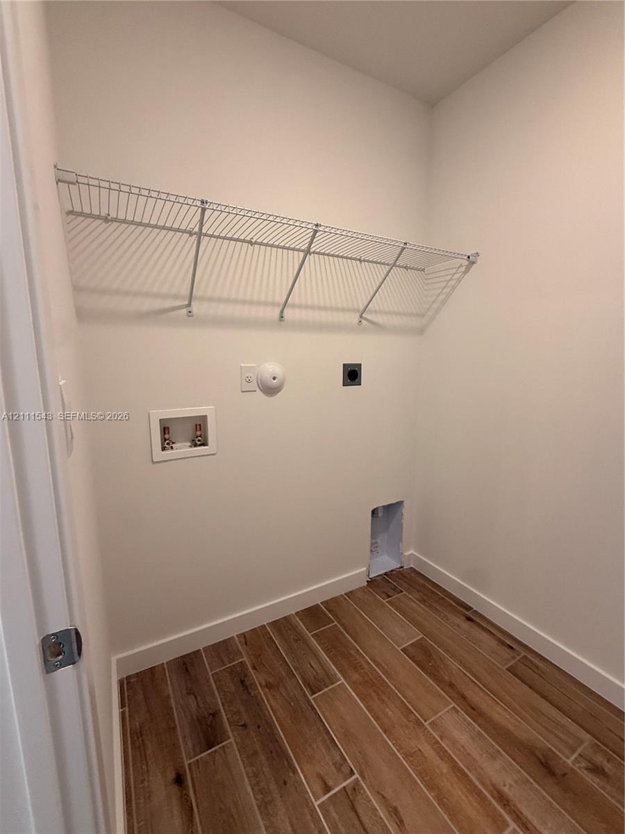 Utility area featuring wood-finish flooring, white walls, and a wall-mounted wire storage shelf, equipped with dedicated water supply lines and laundry drainage hookups