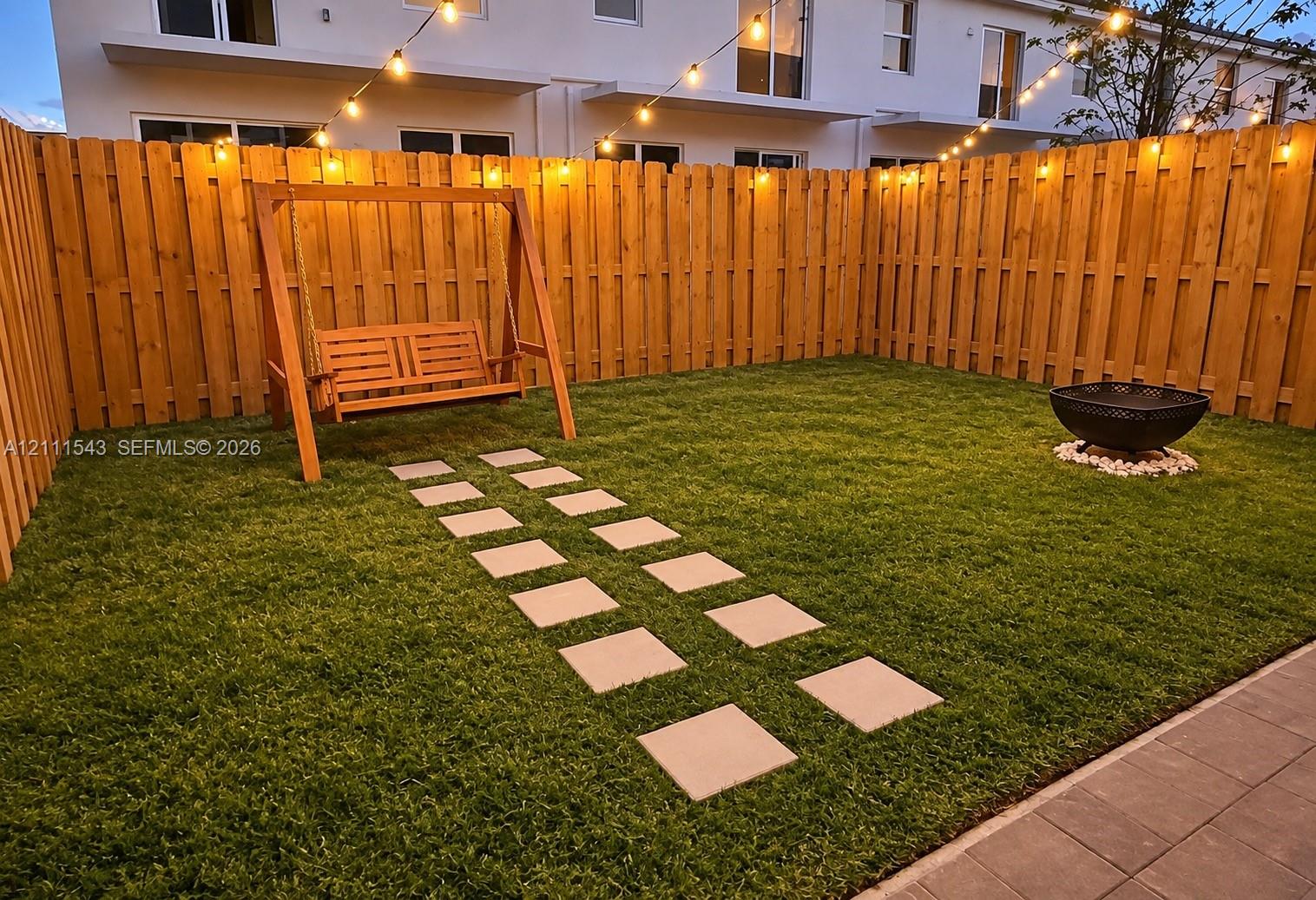 Private backyard featuring a full-height wood privacy fence, synthetic turf lawn, stone paver pathway, and string lighting