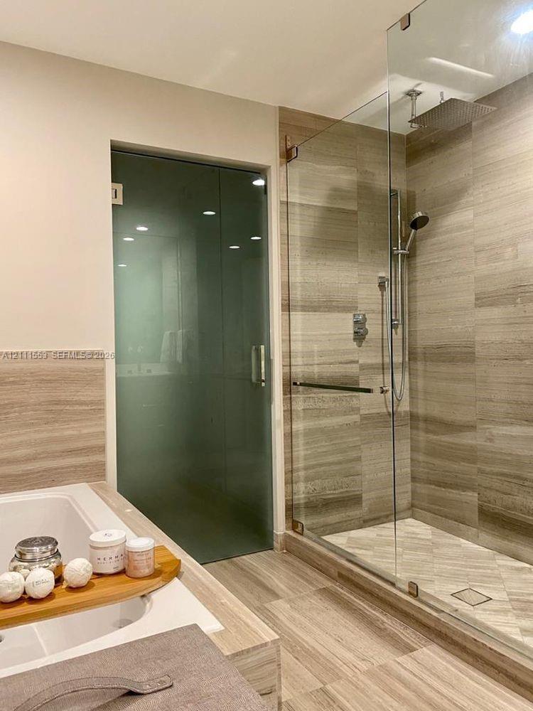 Walk-in glass enclosed shower featuring horizontal wall tile, herringbone floor tile, and a rainfall showerhead, alongside a built-in bathtub surround with stone-finish surfaces and a frosted glass door