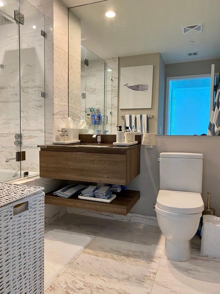 Floating wood-finish vanity with an open lower shelf, glass-enclosed shower, stone-patterned wall tile, and floor tile
