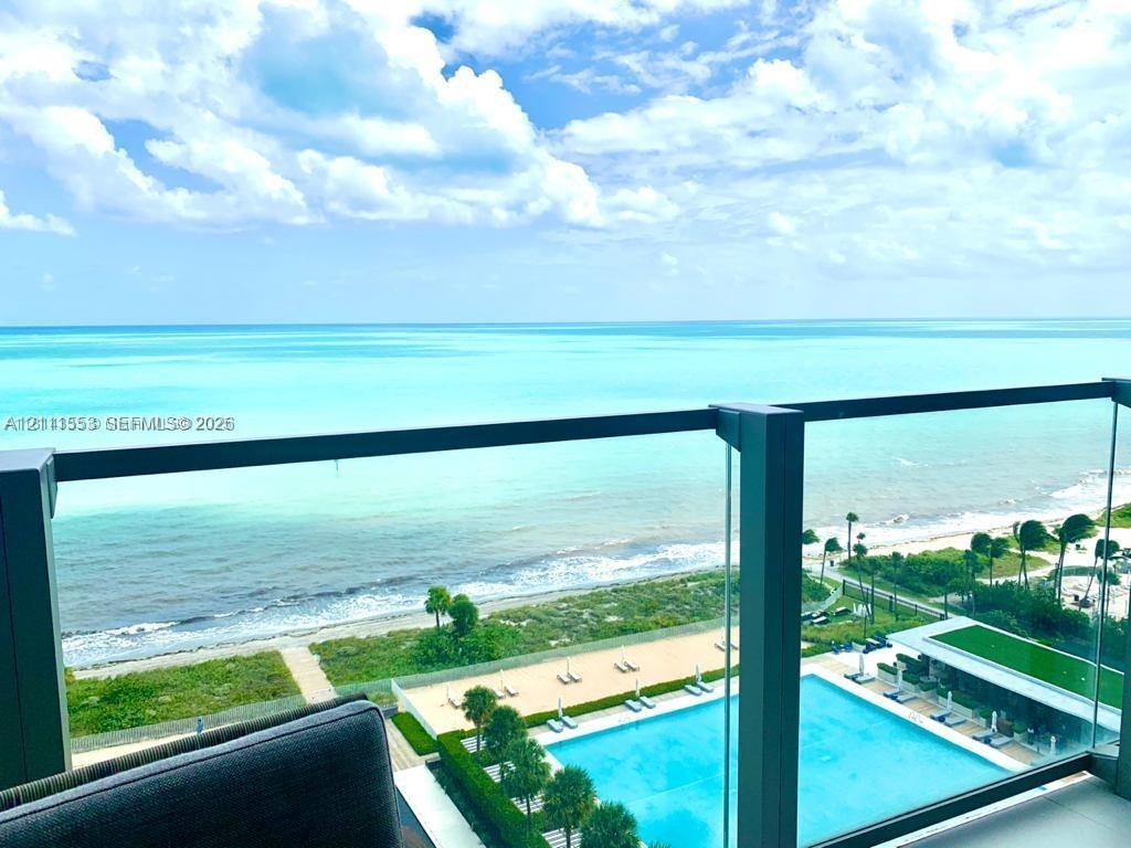 Private balcony featuring a glass-panel railing, overlooking a rectangular swimming pool, manicured grounds, and the ocean