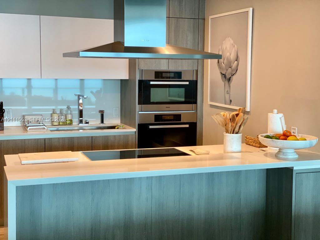 Modern kitchen featuring a waterfall-edge countertop, stainless steel chimney-style range hood, stacked built-in wall ovens, wood-finish cabinetry, and a glass tile backsplash