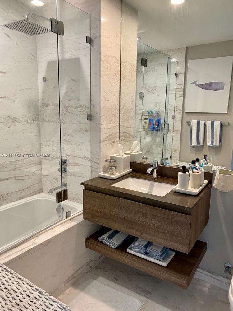 Floating wood-finish vanity with an integrated rectangular basin, large-format stone-patterned wall and floor tile, glass shower enclosure, and square rainfall showerhead