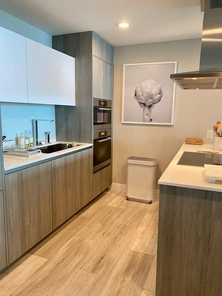 Kitchen featuring flat-panel wood-finish cabinetry, quartz countertops, built-in stainless steel double oven, stainless steel range hood, and wood-finish flooring